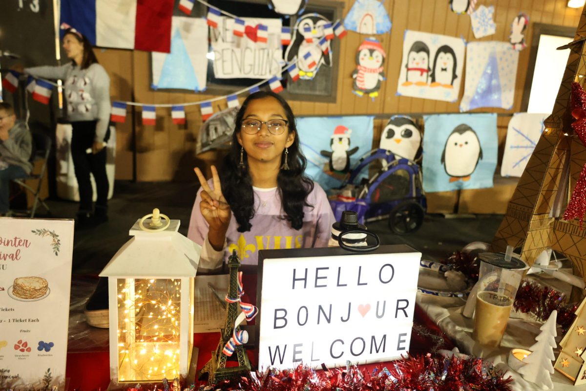 Christa Ashok ('27) poses in front of a festively decorated booth for French Honors Society draped in holiday baubles at the Winter Carnival. 